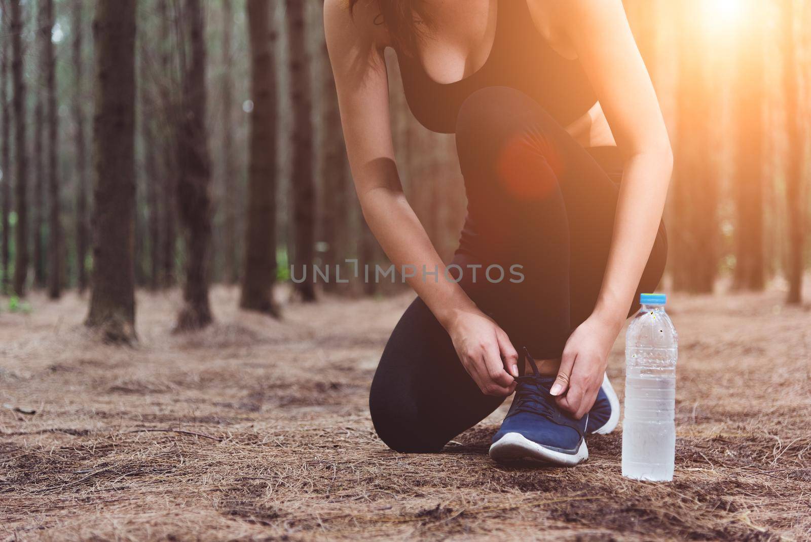 Woman tying up shoelaces when jogging in forest back with drinking water bottle beside hers. Sneakers rope tying. People and lifestyles concept. Healthcare and Wellness theme. Park and Outdoors theme.