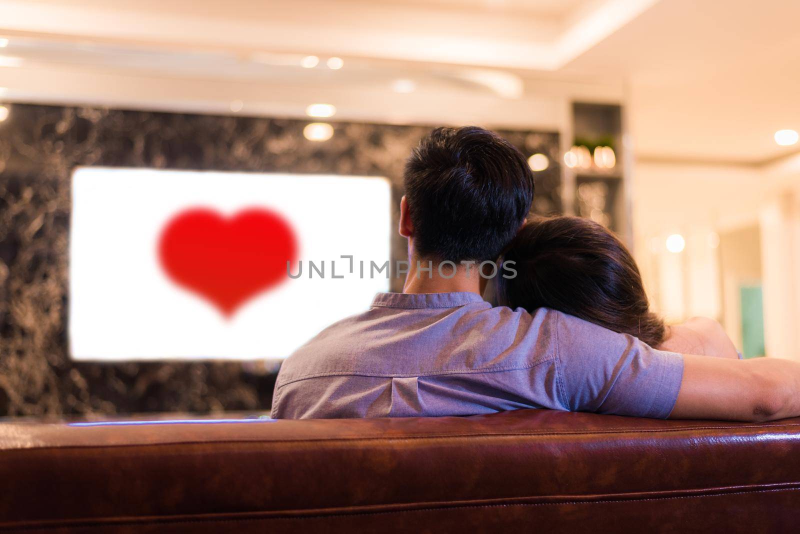 Asian young lovers watching television on sofa. Couples and Relax concept. Holiday and vacation concept. Night dating and honeymoon theme. Back view. Red heart in white screen television