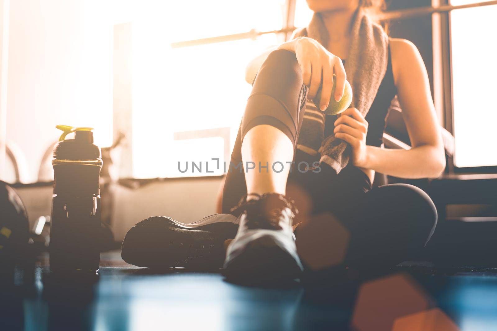 Sport woman sitting and resting after workout or exercise in fitness gym with protein shake or drinking water on floor. Relax concept. Strength training and Body build up theme. Warm and cool tone 