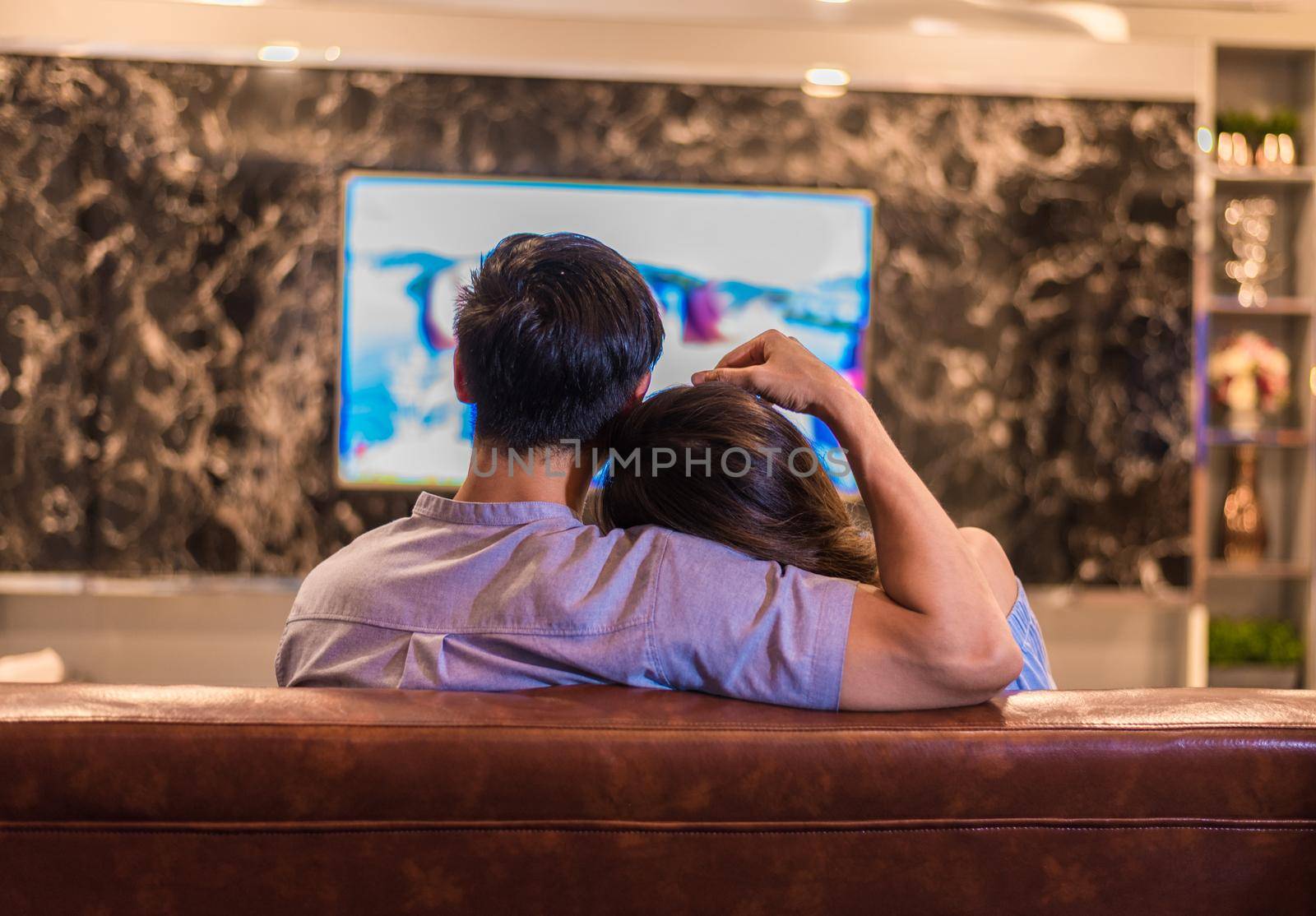 Asian young lovers watching television on sofa. Couples and Relax concept. Holiday and vacation concept. Night dating and honeymoon theme. Back view