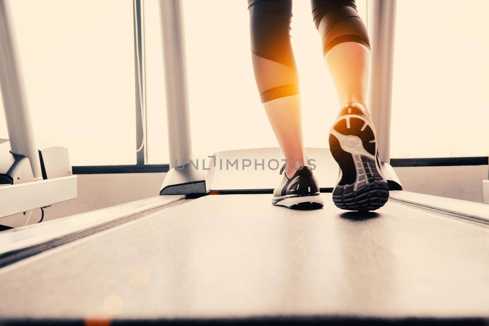 Lower body at legs part of Fitness girl running on running machine or treadmill in fitness gym with sun ray. Warm tone. Healthy and Exercise activity concept. Workout and  Strength training theme.
