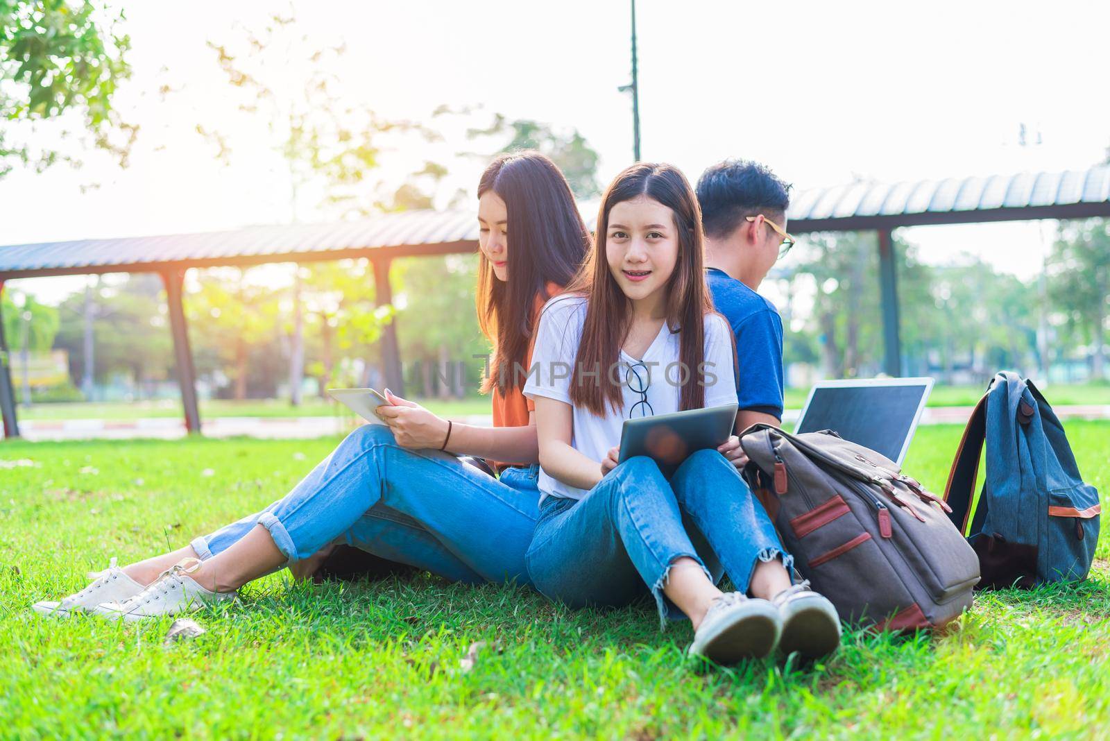 Group of Asian college student using tablet and laptop on grass field at outdoors. Technology and Education learning concept. Future Technology and Modern entertainment concept. Edutainment theme.