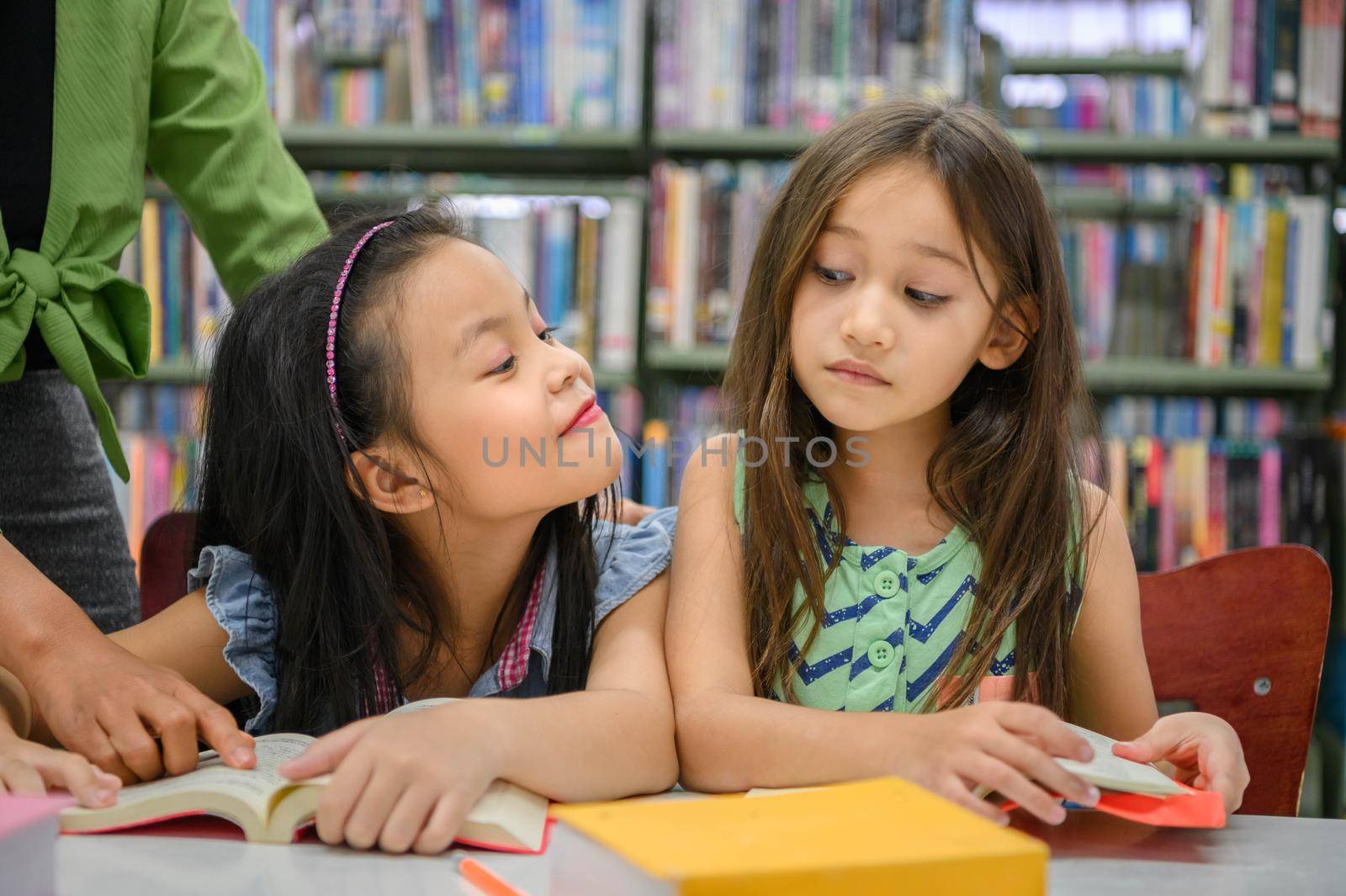 Two cute girls are jealous of each other while reading books in library while teacher teaching. People lifestyles and education. Young friendship and Kids relationship in school concept. Daycare theme