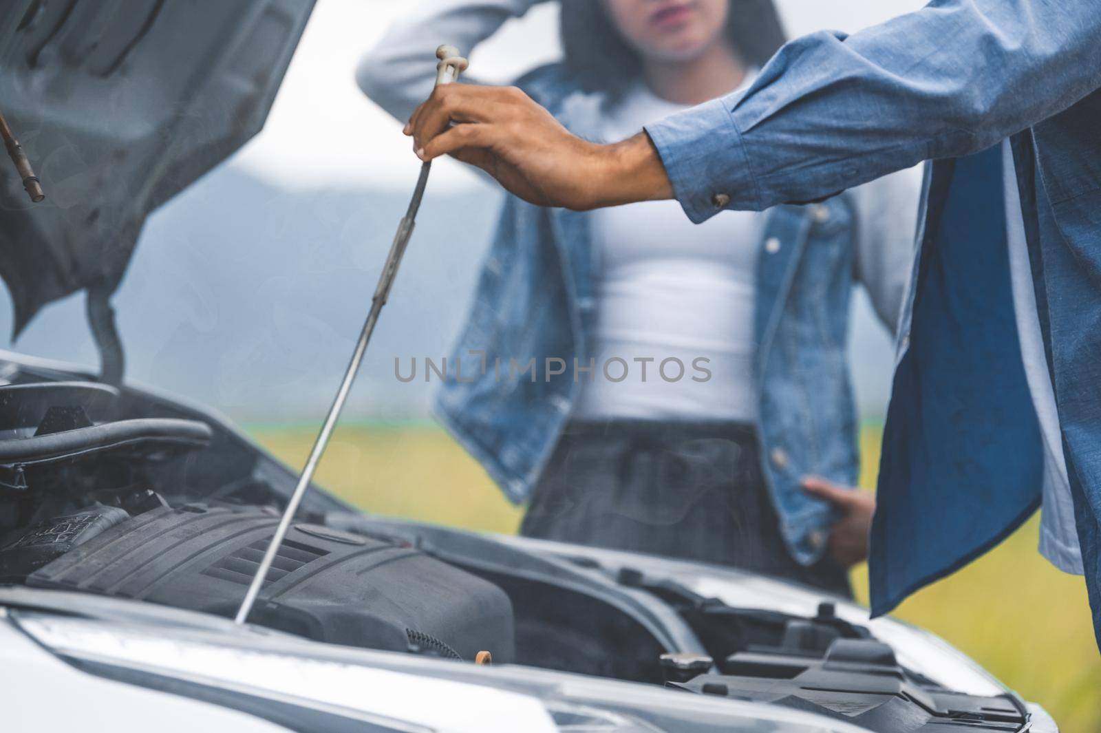 Asian man open car hood for repair as maintenance service with woman background between long road trip. Car broken and breakdown in mountain meadow with wife. People and transport concept