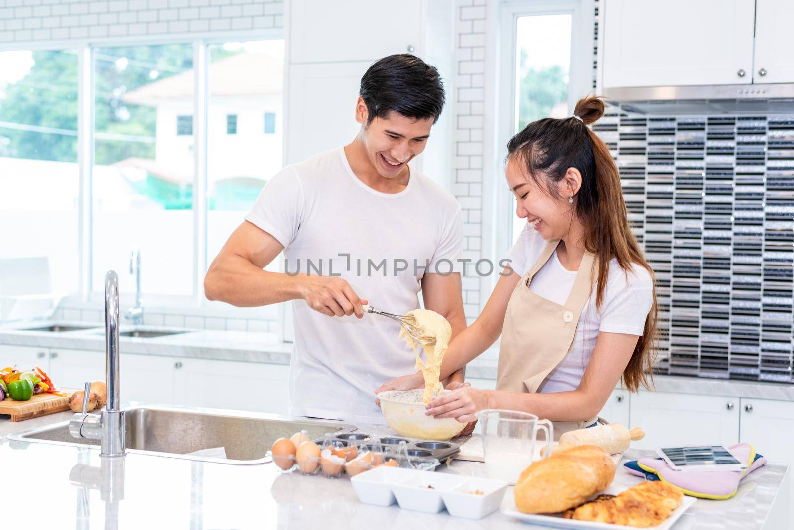 Happy Asian couples cooking and baking cake together in kitchen room. Man and woman looking to tablet follow recipe step at home. Love and happiness concept. Sweet honeymoon and Valentine day theme