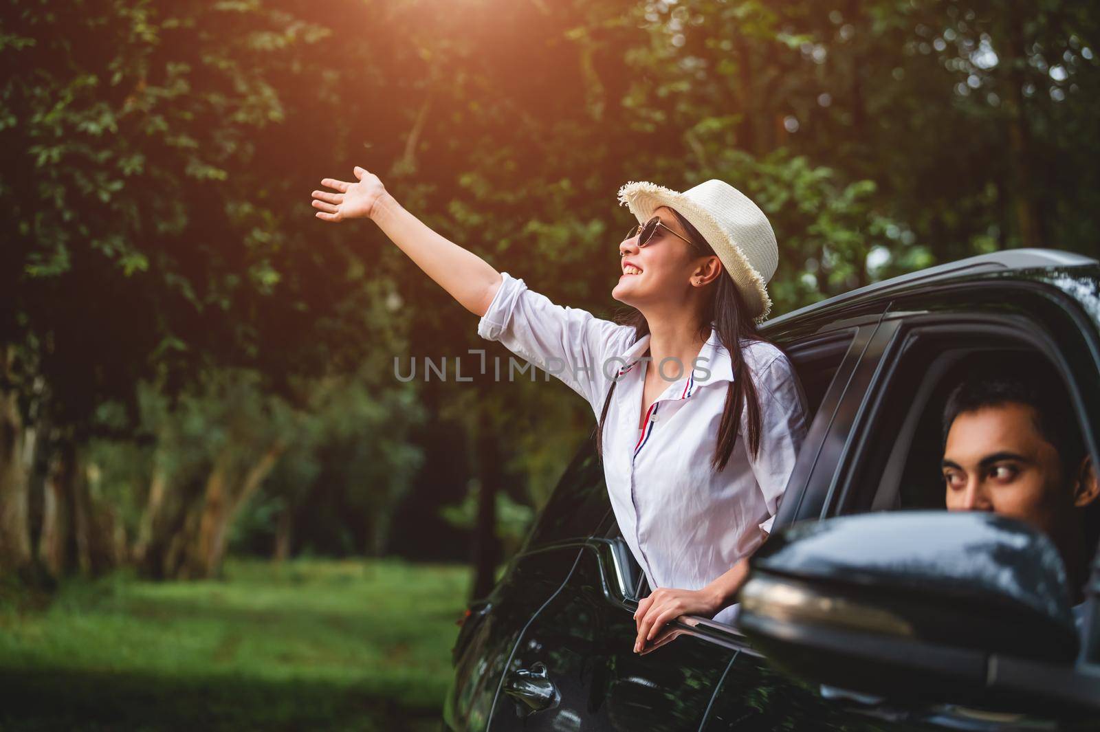 Happy woman waving hand outside open window car with her boyfriend on forest background. People lifestyle relaxing as traveler on road trip in holiday vacation. Transportation and weekend travel