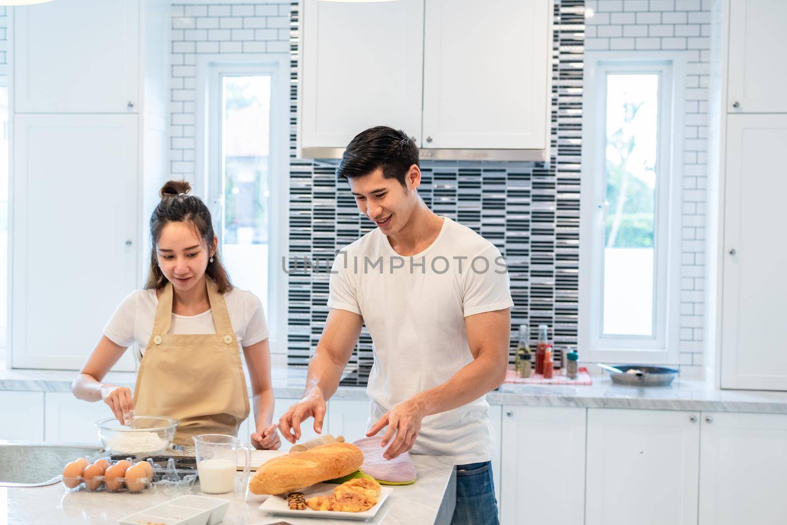 Happy Asian couples cooking and baking cake together in kitchen room. Man and woman looking to tablet follow recipe step at home. Love and happiness concept. Sweet honeymoon and Valentine day theme