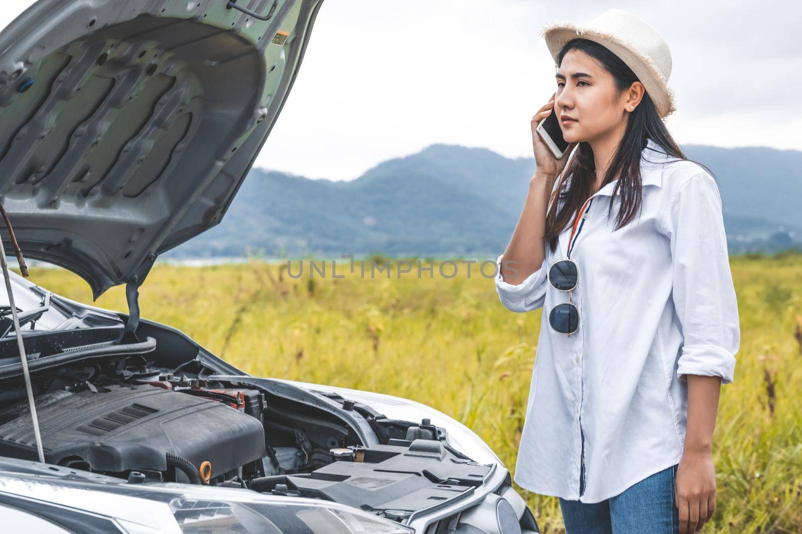 Asian woman calling car mechanic service for repairing breakdown broken car by mobile phone during driving to destination. Car maintenance and transportation concept. People travel and landscape theme