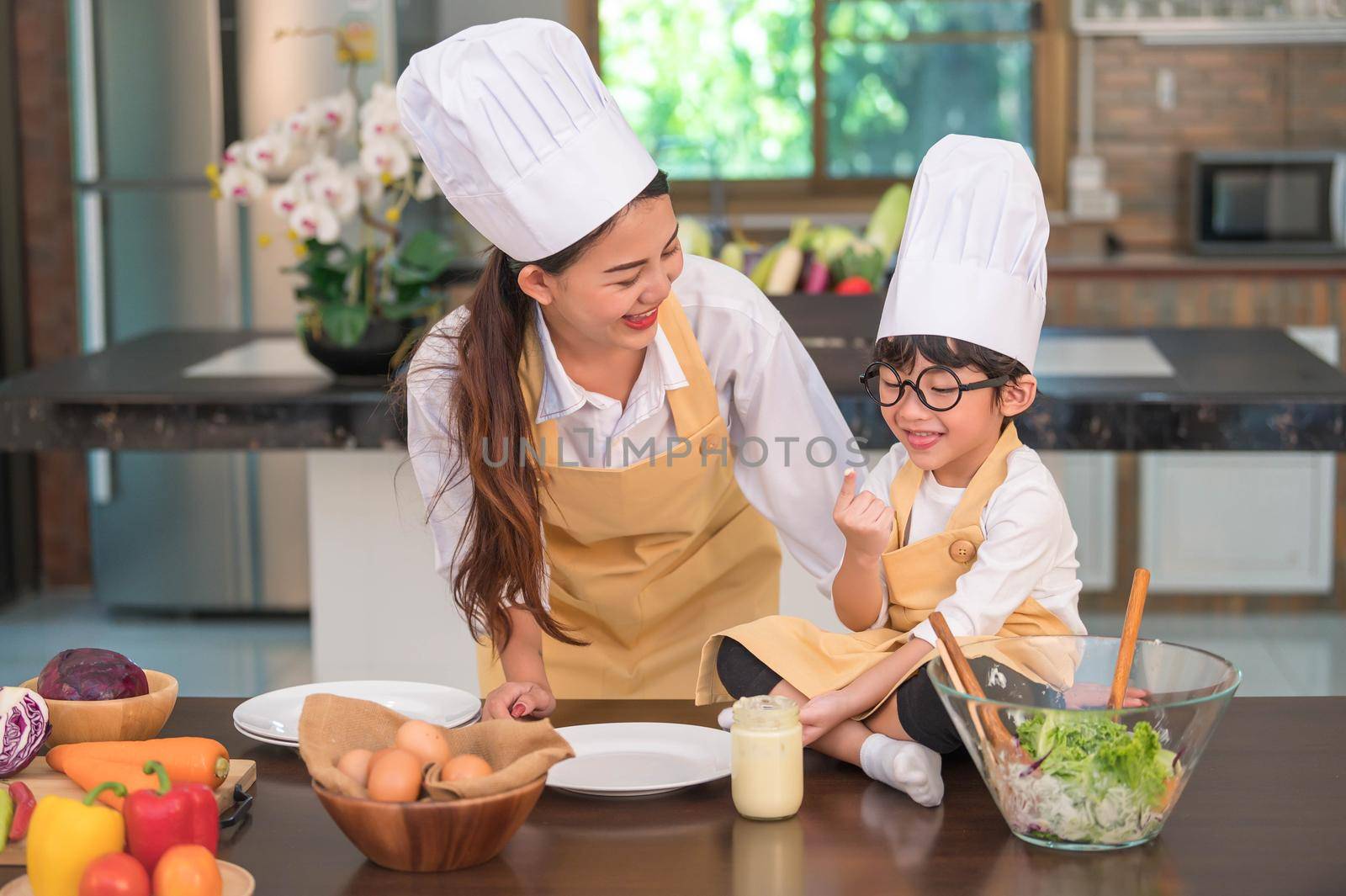 Happy beautiful Asian woman and cute little boy with eyeglasses prepare to cooking in kitchen at home. People and Family concept. Homemade food and ingredients concept. Two Thai people lifestyles