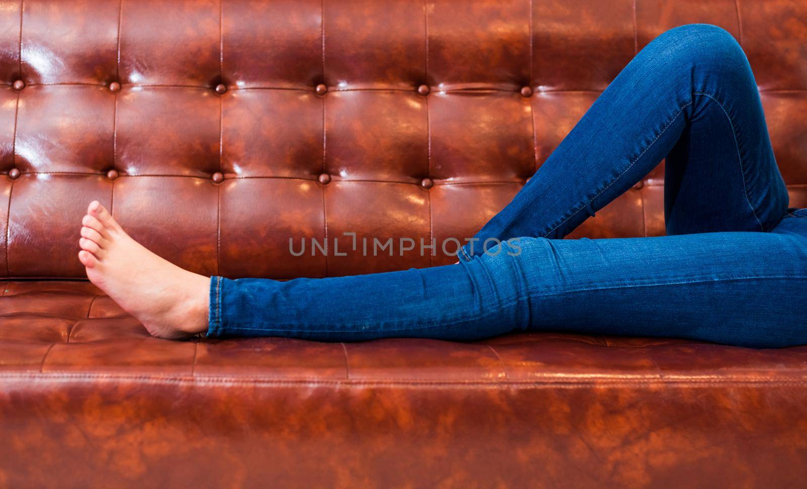 Woman legs with wearing jeans while relaxing at movie theater or home on brown leather sofa, Relax and holiday concept, Bed time sleeping concept. People lifestyle and holiday. Closeup girl foot