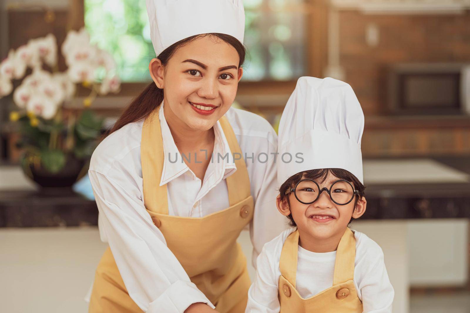 Portrait cute little Asian boy chef with eyeglasses and his mother looking to camera in home cooking kitchen happily and funny. People lifestyles and Family. Homemade food ingredient making concept