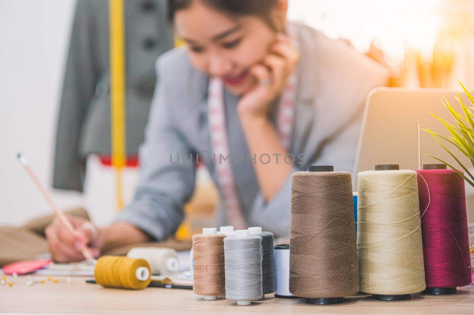 Closeup of color thread with blurry happy female tailor and sewing clothing stylist designer background. Woman creating new collection in workshop office. People lifestyle and freelance occupation job