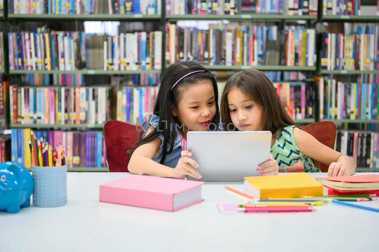 Two little happy cute girls playing on a tablet PC computing device in library at school. Education and self learning wireless technology concept. People lifestyles and friendship. Preschool children