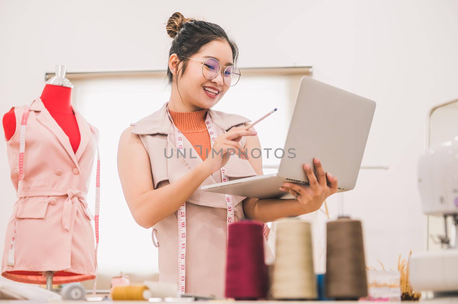 Happy Asian female fashion designer working in clothing shop studio and using laptop computer to contact customer. Woman creating design new collection . Tailor sewing. People lifestyle concept