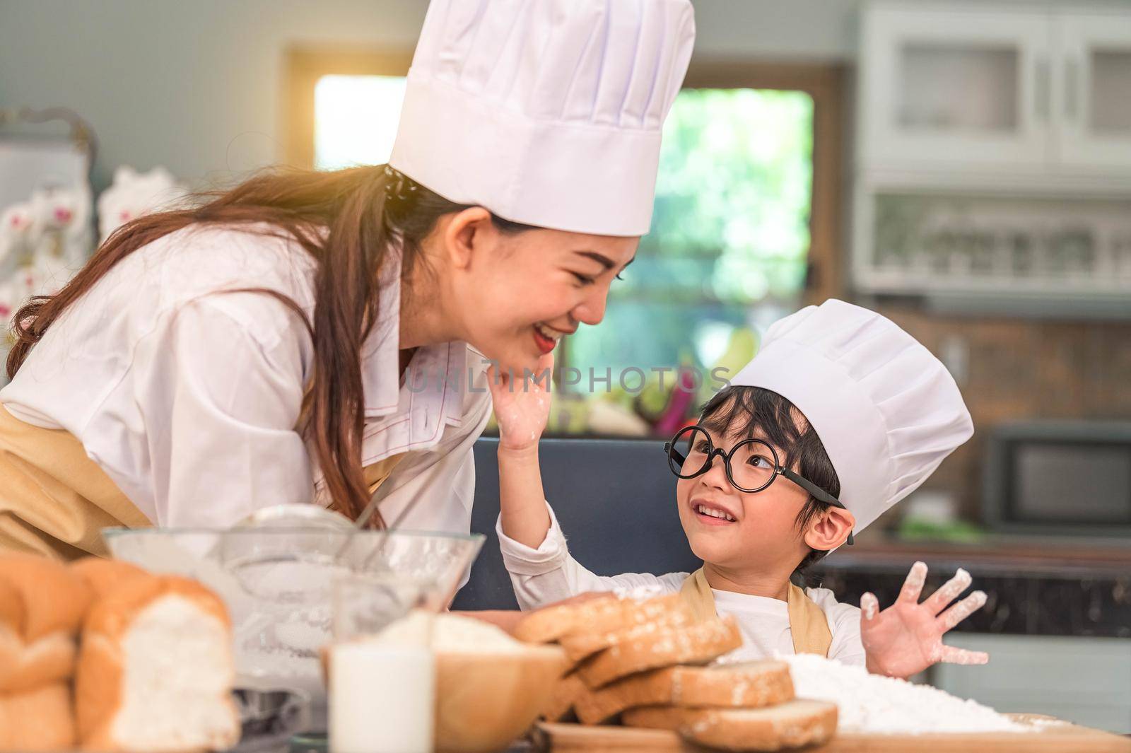 Cute little Asian boy painting beautiful woman face with dough flour. Chef team playing and baking bakery in home kitchen funny. Homemade food and bread. Education teamwork and learning concept.