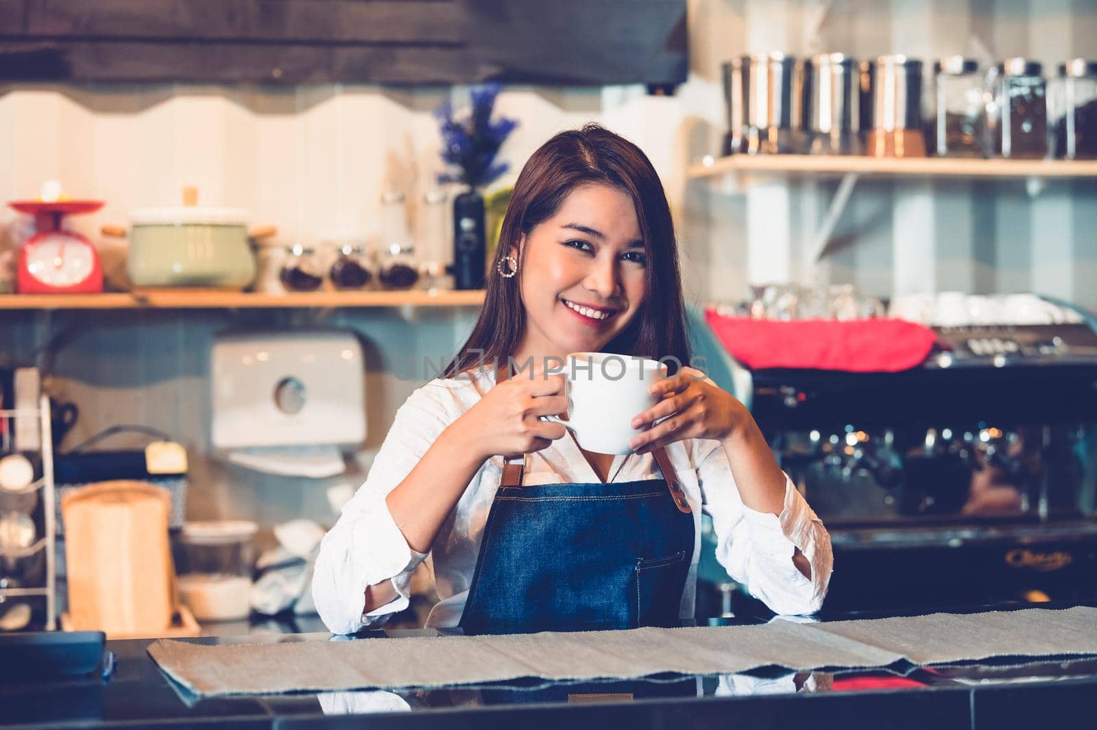 Asian female barista making cup of coffee. Young woman holding white coffee cup while standing behind cafe counter bar in restaurant background. People lifestyles and Business occupation concept.