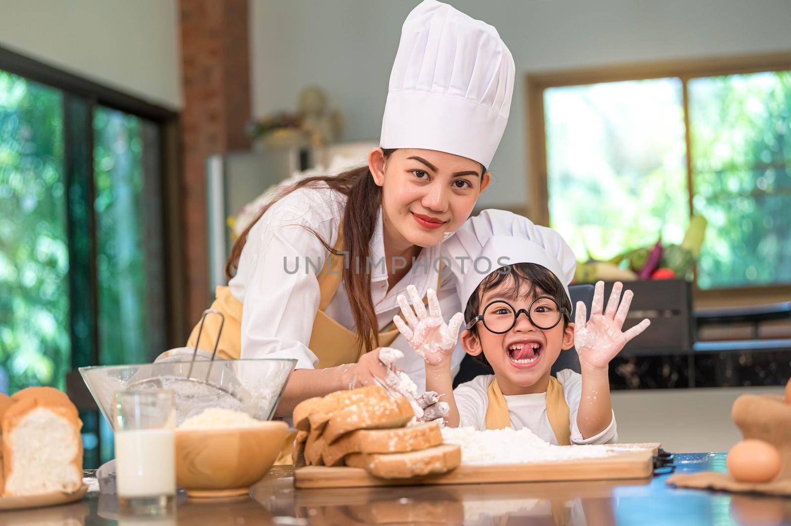 Beautiful woman and cute little Asian boy with eyeglasses, chef hat and apron playing and baking bakery in home kitchen funny concept. Homemade food and bread. Funny face emotion looking camera