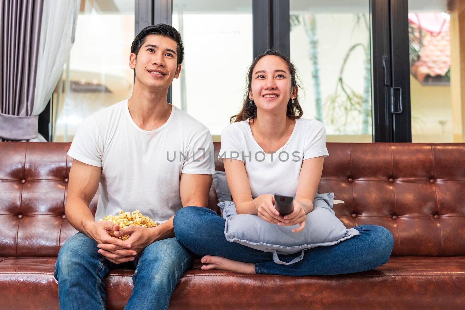 Asian couples watching television together on sofa in their home. People and lifestyles concept. Vacation and holiday concept. Honeymoon and pre wedding theme. Happy family activity in valentines day by MiniStocker