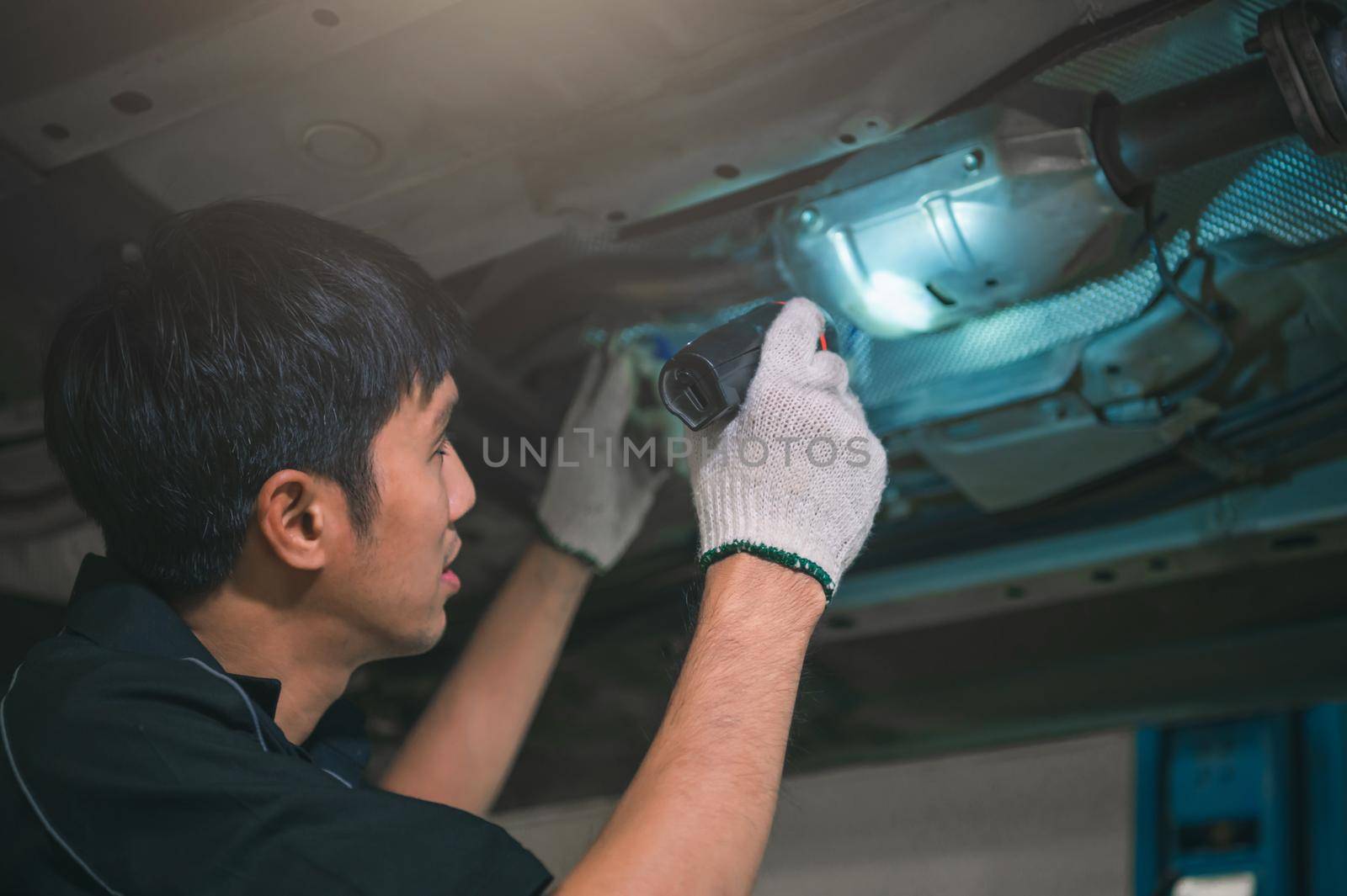 Asian male mechanical hold and shining flashlight to examine car under chassis of automotive vehicle. Safety suspension inspection check service maintenance for customer before road trip concept