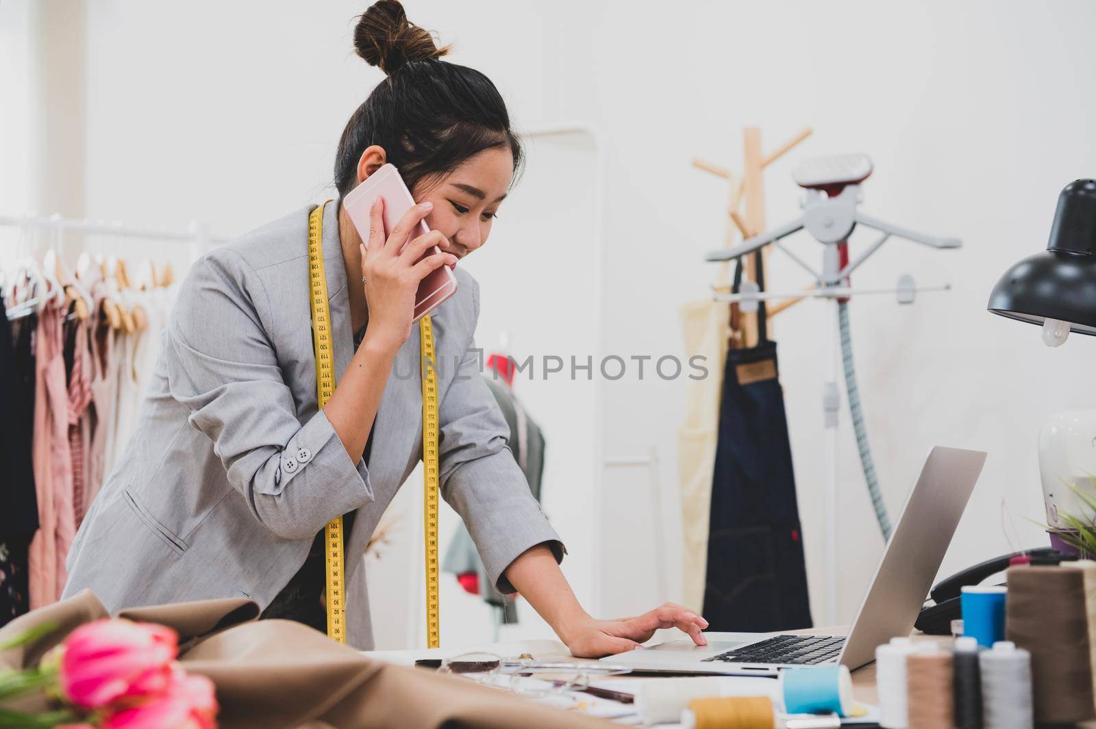 Attractive Asian female fashion designer working in clothing shop studio and using mobile phone to contact customer. Woman receiving order from incoming call. Tailor sewing. People lifestyle concept