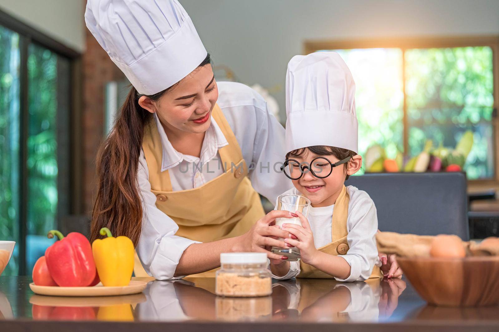 Asian mother helping cute little boy drinking milk in glass at home kitchen in chef cooking uniform. People lifestyles and Happy family togetherness concept. Calcium and protein in milk nutrition