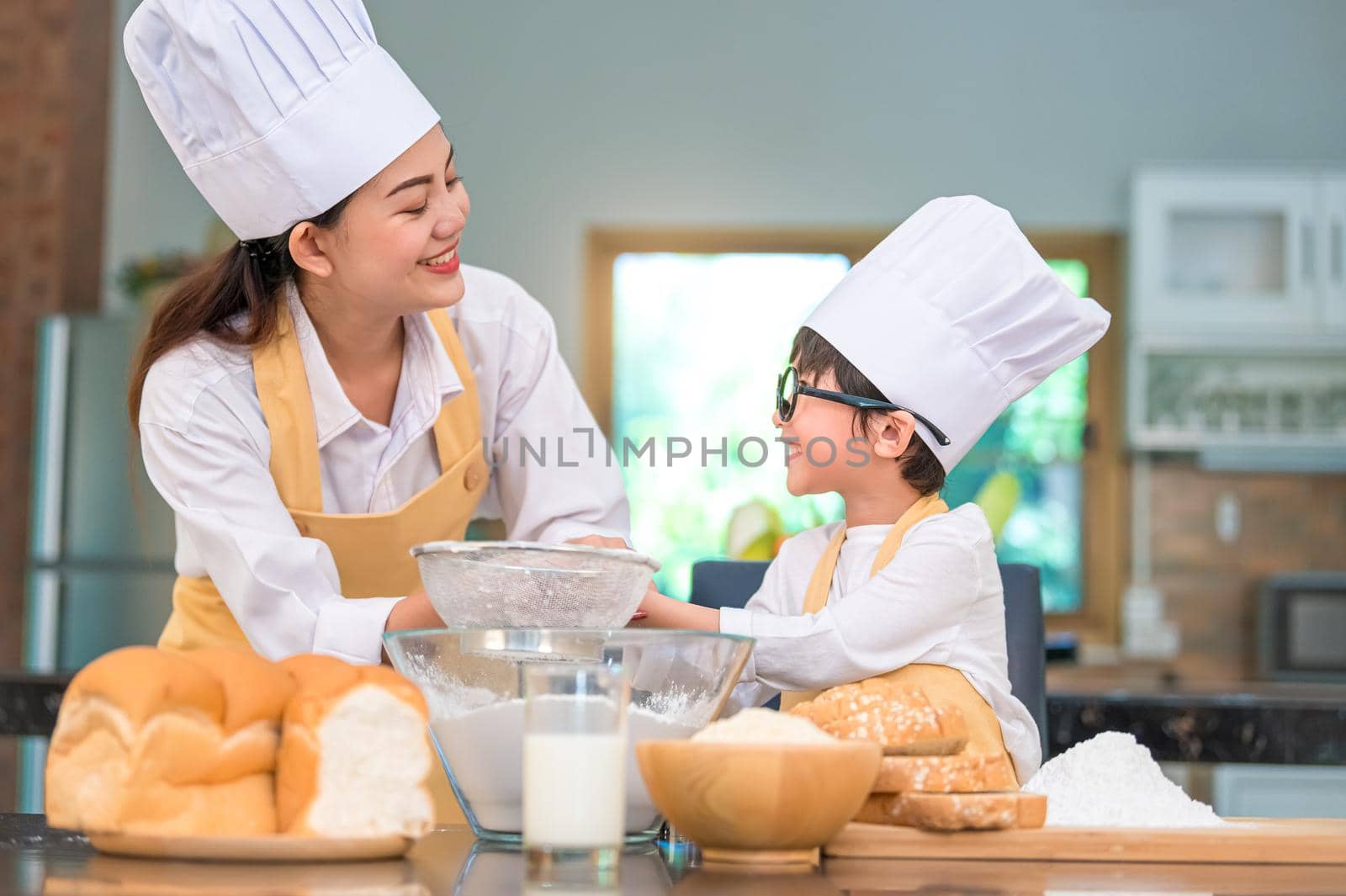 Cute little Asian boy and beautiful mother sifting dough flour with sifter sieve colander in home kitchen on table for prepare to baking bakery and cake. Thai kids playing with flour as chef funny