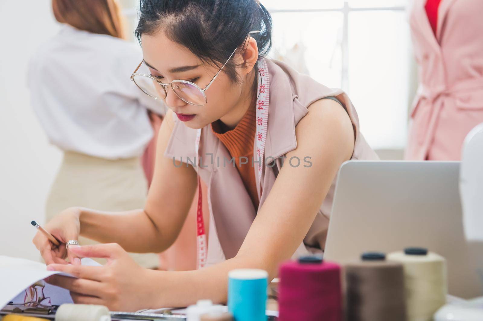 Attractive Asian female fashion designer working in clothing shop with customer background. Stylish fashionista woman creating new cloth design collection. Tailor and sewing. People lifestyle concept