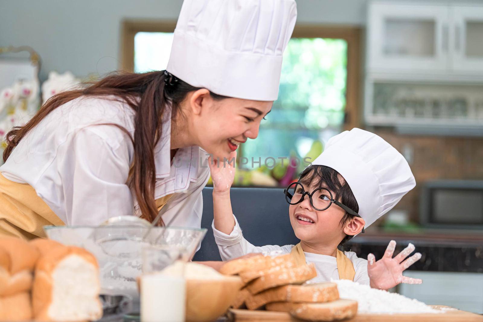 Cute little Asian boy painting beautiful woman face with dough flour. Chef team playing and baking bakery in home kitchen funny. Homemade food and bread. Education teamwork and learning concept.