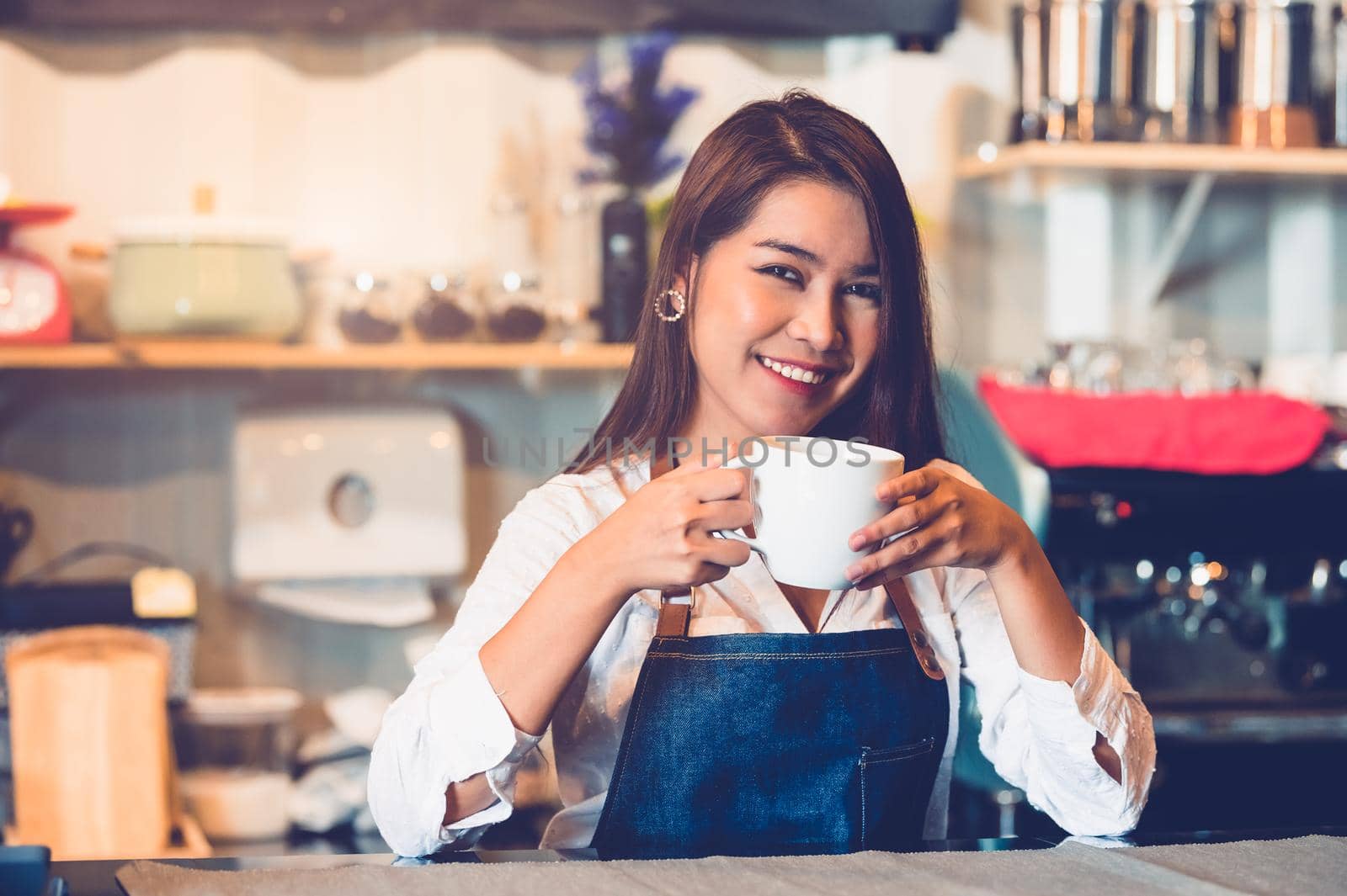 Asian female barista making cup of coffee. Young woman holding white coffee cup while standing behind cafe counter bar in restaurant background. People lifestyles and Business occupation concept.