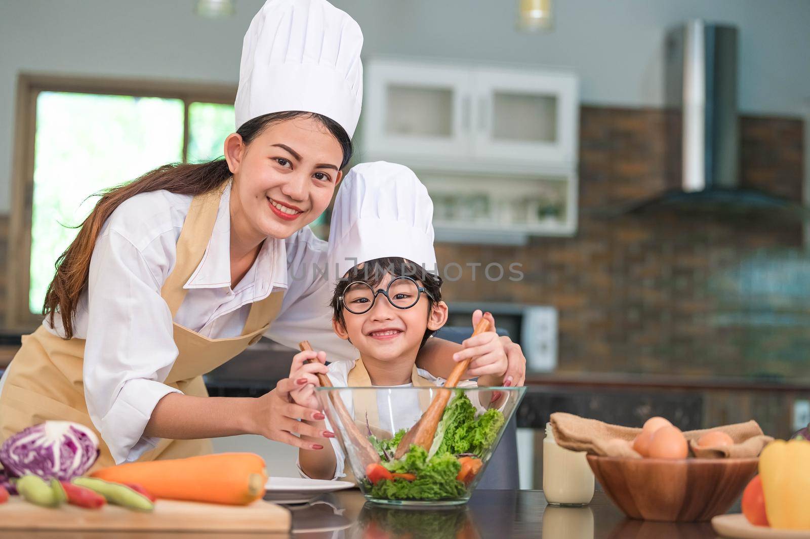 Beautiful Asian woman and cute little boy with eyeglasses prepare to cooking in kitchen at home. People lifestyles and Family. Homemade food and ingredients concept. Two Thai people looking at camera
