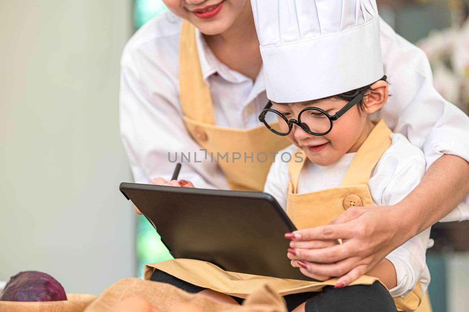 Beautiful Asian woman and cute little boy prepare online shopping and listing ingredient for cooking in kitchen at home with tablet. People lifestyles and Family. Homemade food and ingredient concept.