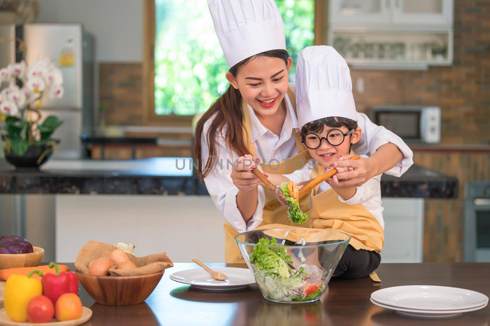 Happy beautiful Asian woman and cute little boy with eyeglasses prepare to cooking in kitchen at home. People lifestyles and Family. Homemade food and ingredients concept. Two Thai people life