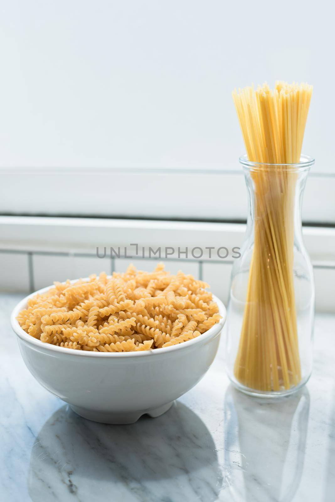 White bowl of spiral macaroni and tied spaghetti stand erect at windows of kitchen room. Food and ingredients concept. Italian food theme
