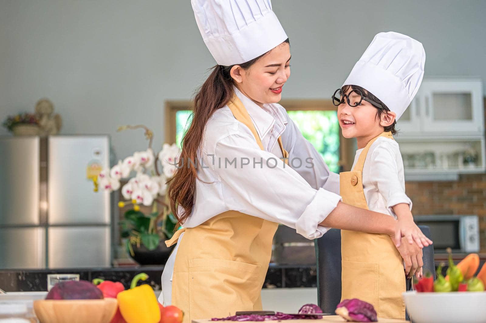 Happy beautiful Asian woman dress up cute little boy chef outfit for prepare to cooking in home kitchen. People lifestyles and Family. Homemade food and ingredients concept. Two Thai people life