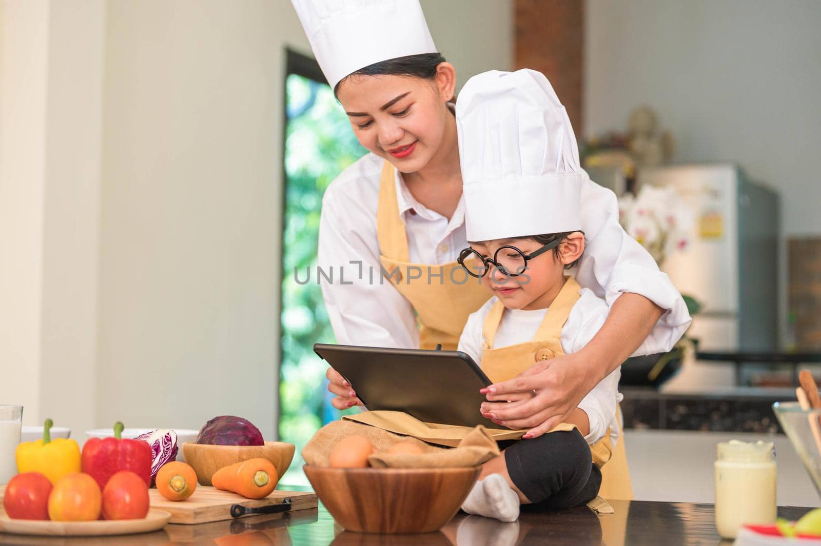 Beautiful Asian woman and cute little boy with eyeglasses prepare to cooking in kitchen at home with tablet. People lifestyles and Family. Homemade food and ingredients concept. Ketogenic salad making