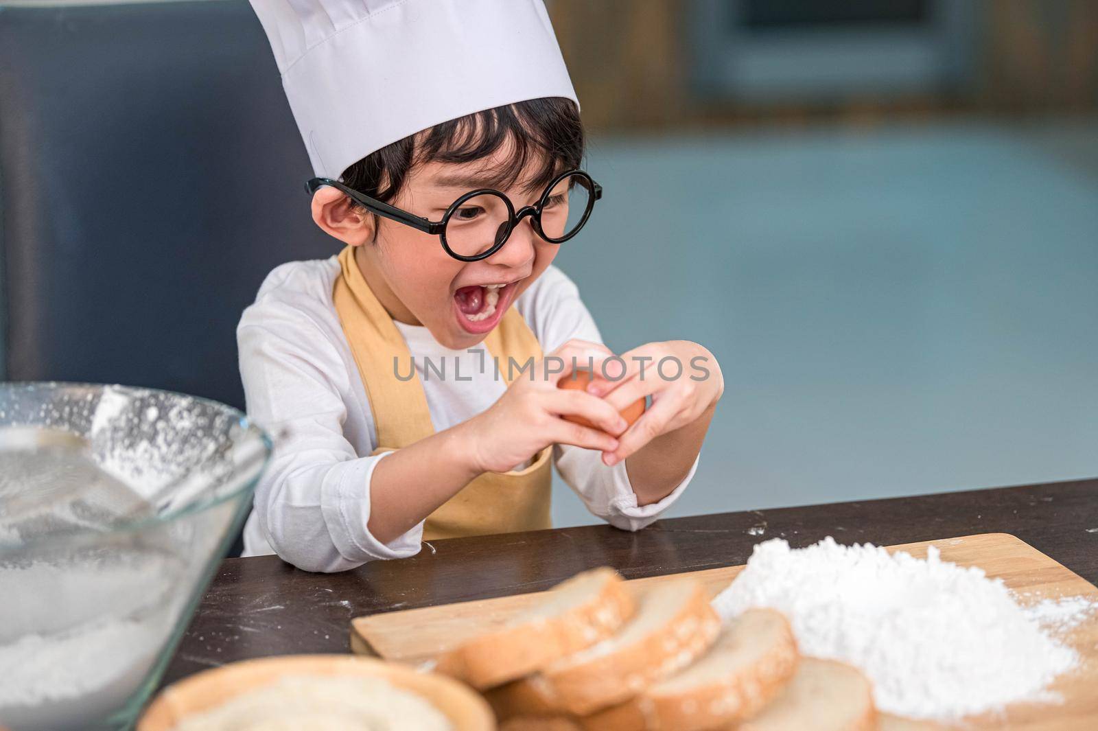 Portrait cute little Asian happy boy surprised and interested in how to break or crack egg cooking funny in home kitchen. People lifestyle and Family. Homemade food learning education and ingredient