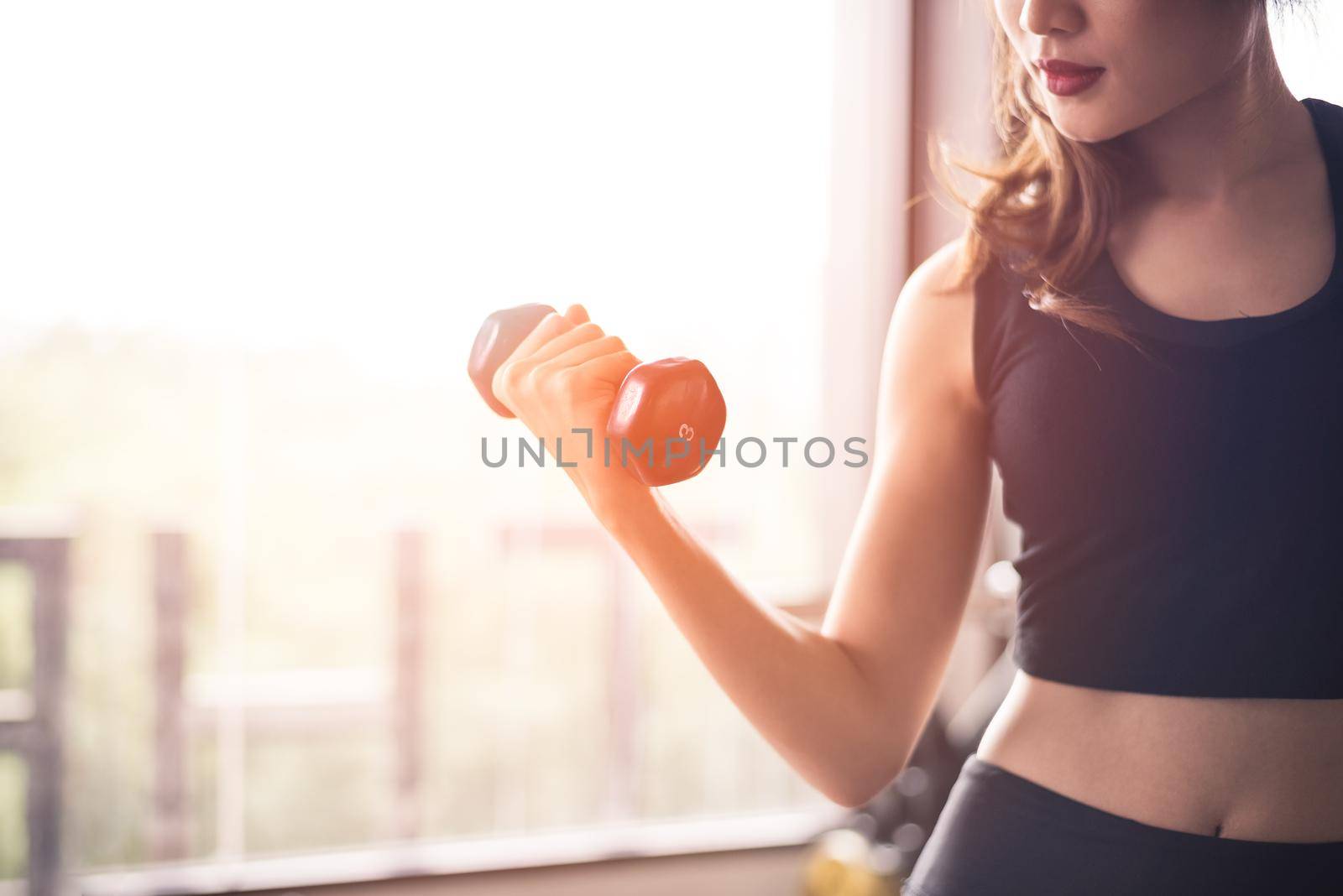 Hand of sports woman lifting dumbbell for weight training near window by right hand for pumping biceps muscle. Workout and Body build up concept. Indoor exercise gym and Healthcare theme