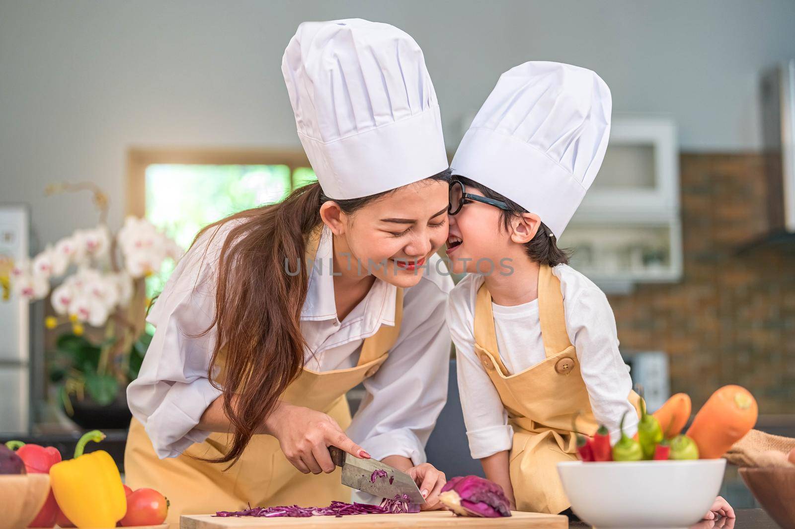Happy cute little boy with eyeglasses kissing beautiful Asian woman mother while prepare to cooking in kitchen at home. People lifestyles and Family. Homemade food and ingredients concept.