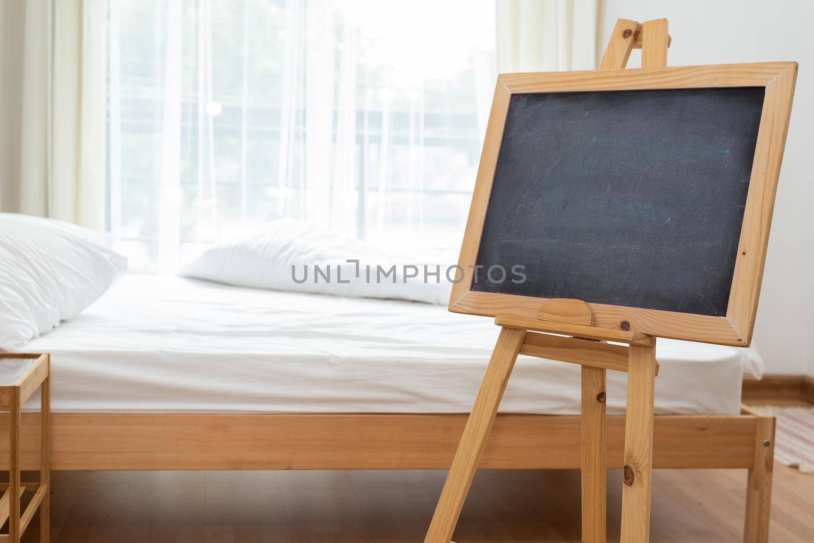 Black chalkboard on easel stand in bedroom with white soft bed background. Interior and decoration concept.