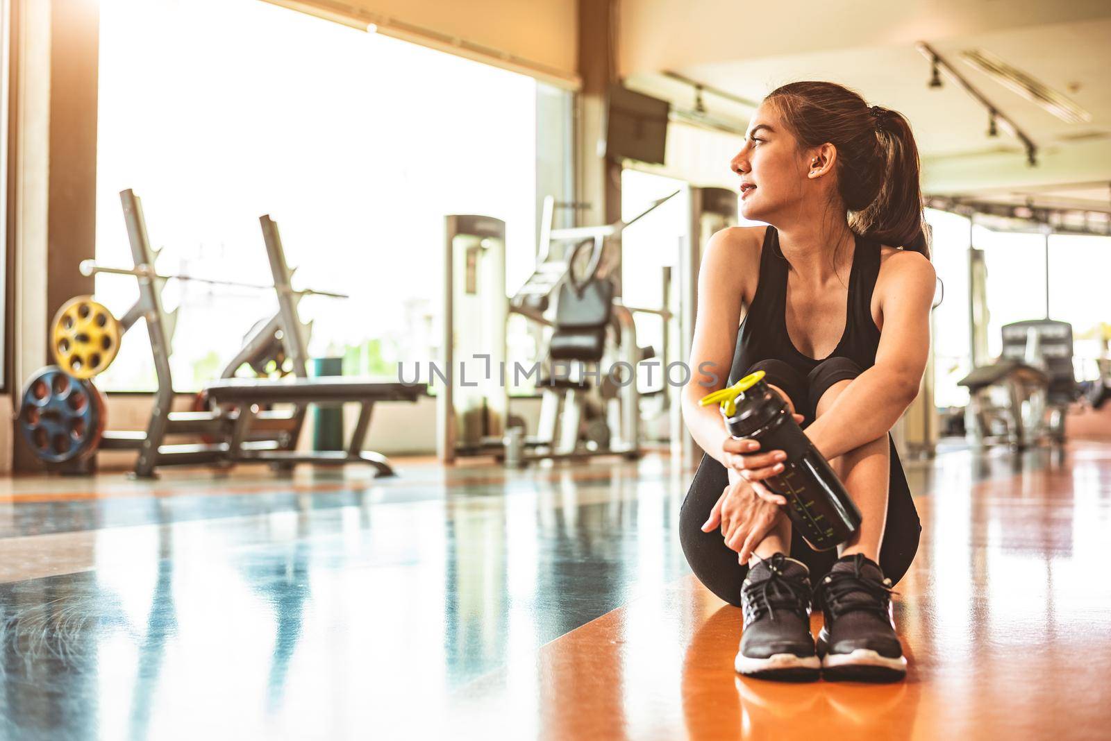 Sport woman relax resting after workout or exercise in fitness gym. Sitting and drinking protein shake or drinking water on floor. Strength training and Bodybuilder muscle theme. Warm and cool tone 