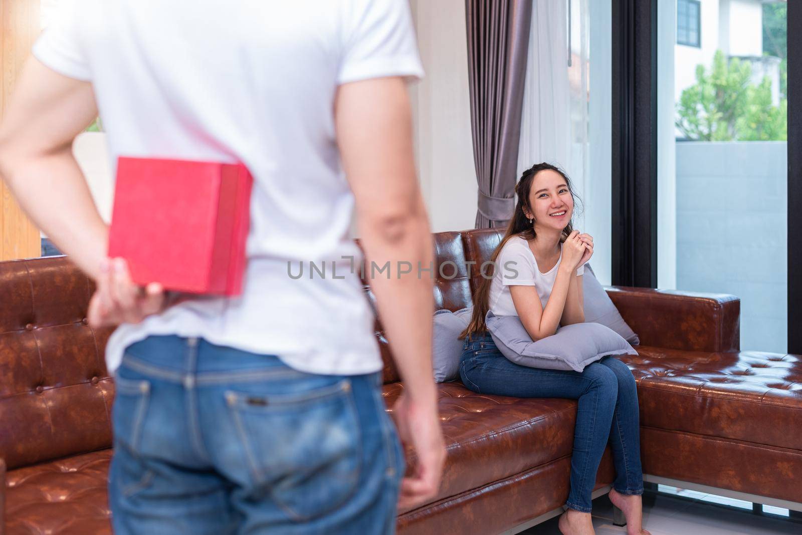 Boy surprise his Asian girlfriend by holding gift box behind him at their home. Valentine's day and pre wedding honeymoon concept. People and lifestyles concept.