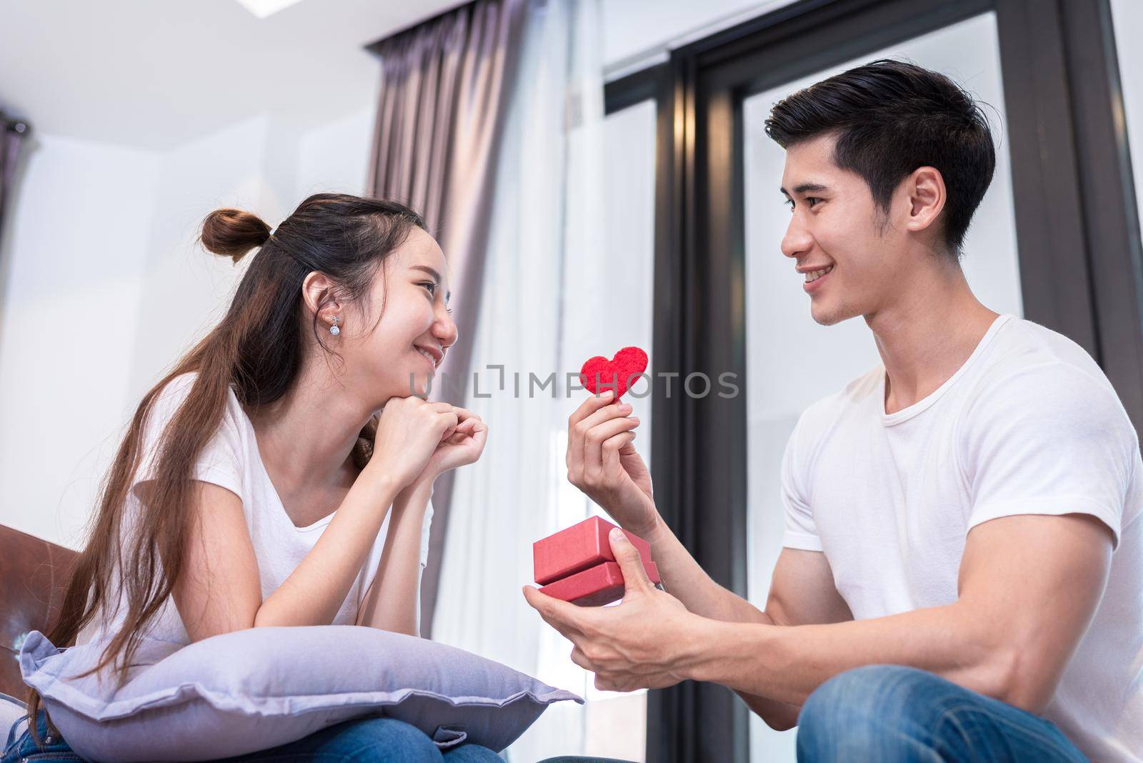 Man holding surprise box and red heart gift to woman on sofa at home in living room Relax and Holiday concept. Happy sweet romantic of young couple in home theme by MiniStocker