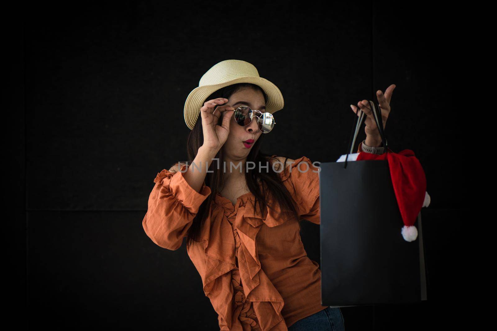 Asian shopping woman surprised with Black Friday shopping bag and Santa Claus hat inside on black background. Shopaholics and beauty fashion theme.
