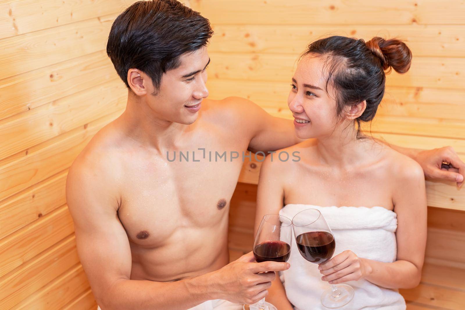 Asian couples toasting and drinking wine in sauna room for celebrating wedding anniversary. Healthy skin heat treatment with steam sauna concept. Holiday and Relax. People lifestyles together