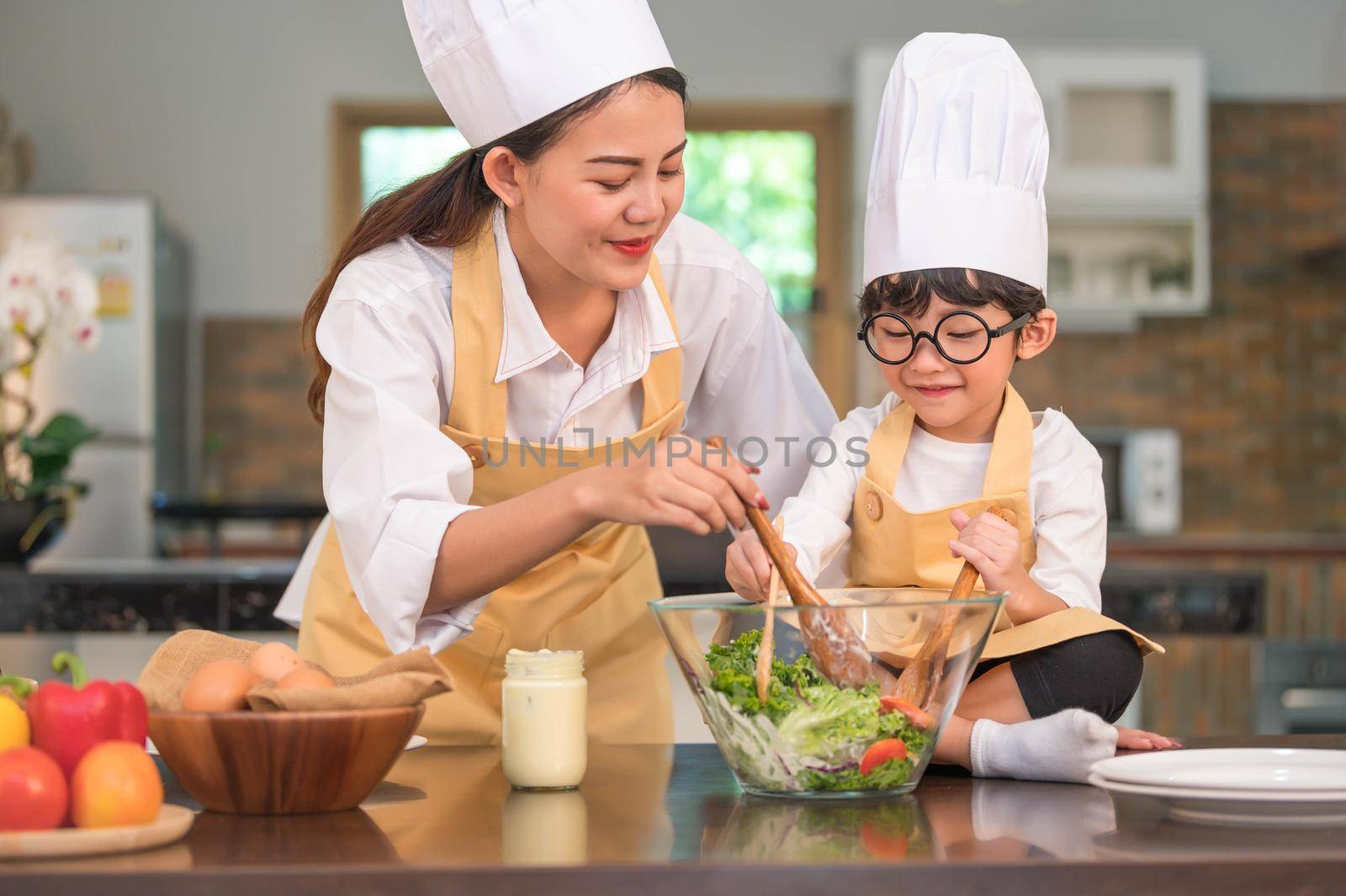 Happy beautiful Asian woman and cute little boy with eyeglasses prepare to cooking in kitchen at home. People lifestyles and Family. Homemade food and ingredients concept. Two Thai people life