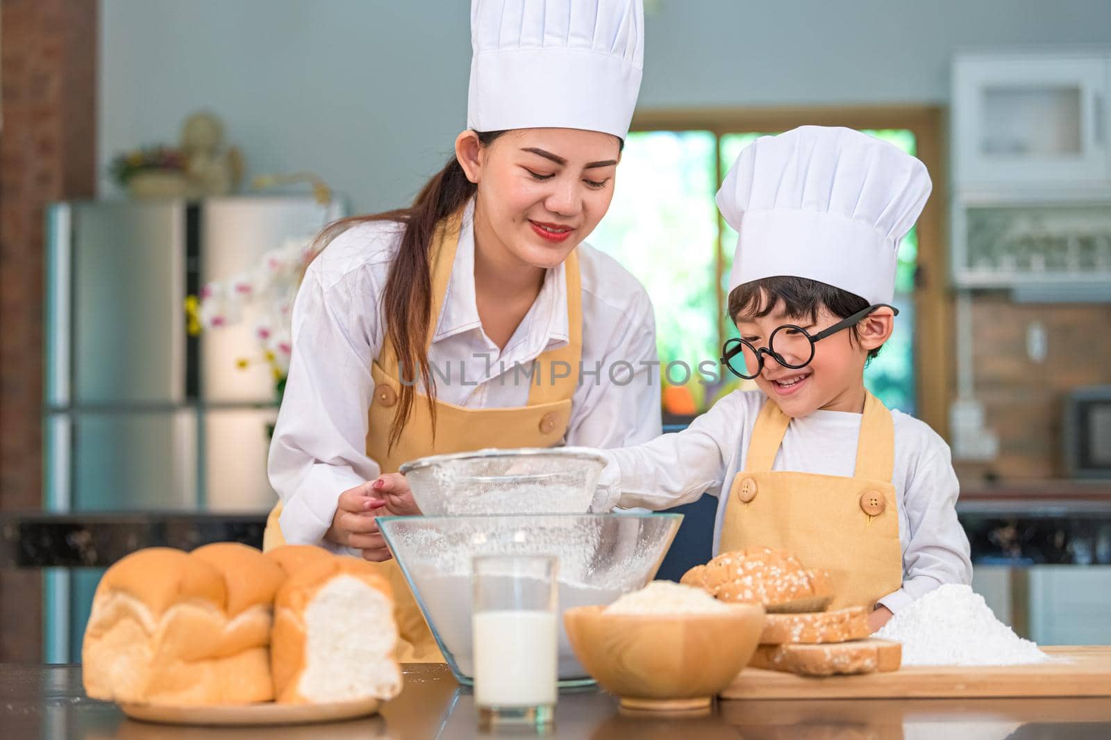 Cute little Asian boy and beautiful mother sifting dough flour with sifter sieve colander in home kitchen on table for prepare to baking bakery and cake. Thai kids playing with flour as chef funny