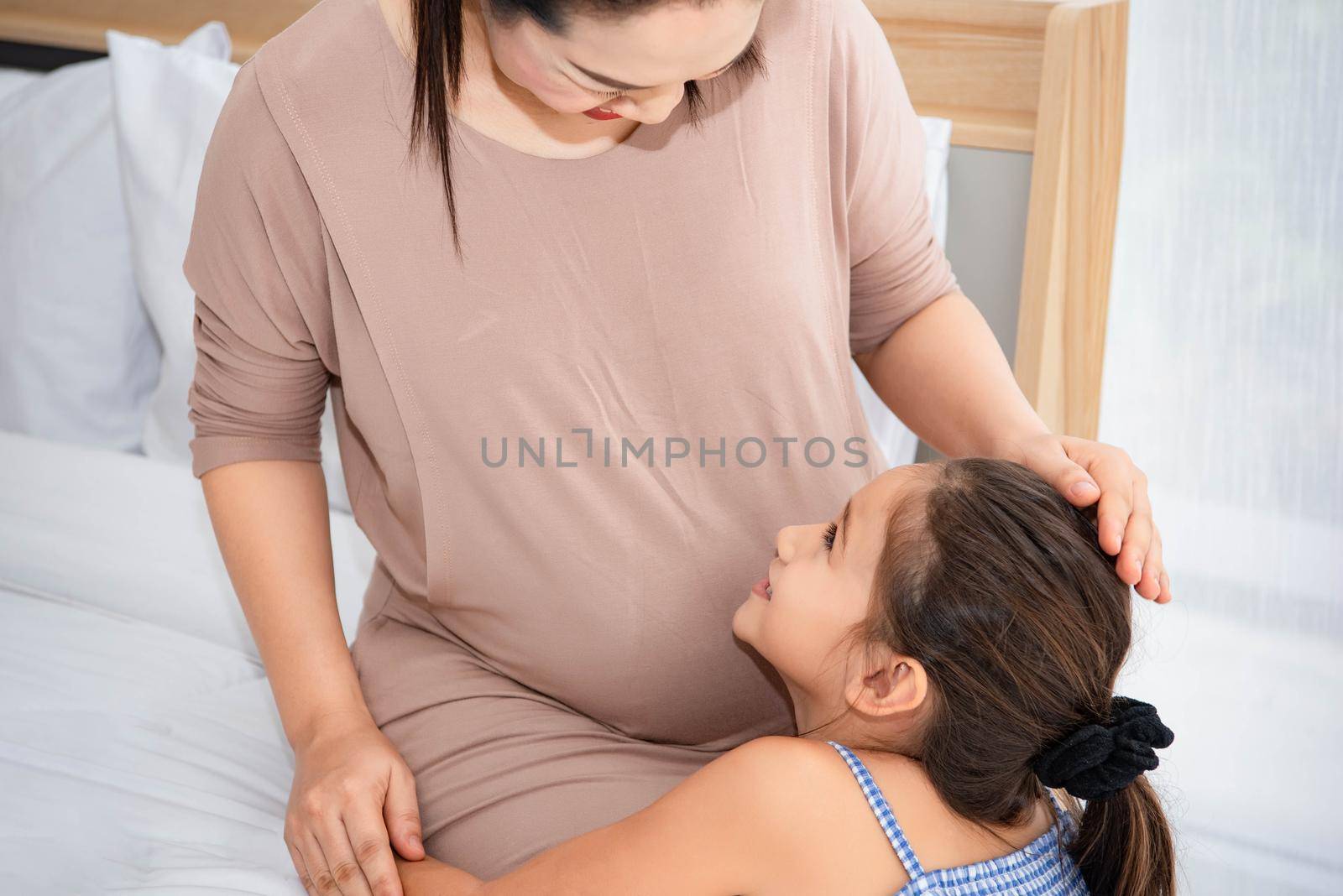 Cute daughter hugging mother and listening to voice of baby in mother belly for checking and looking at mom face. Pregnant and parents. Health and medical. People lifestyles and family love concept. by MiniStocker