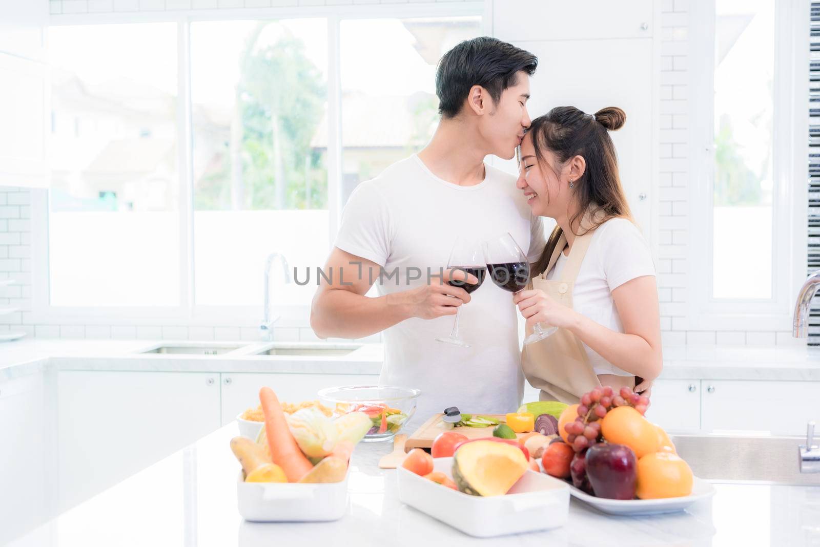 Asian lovers in kitchen, Man give forehead kiss to woman while clink wine glasses to each other. Family and Couple concept. Honeymoon and Holidays theme by MiniStocker