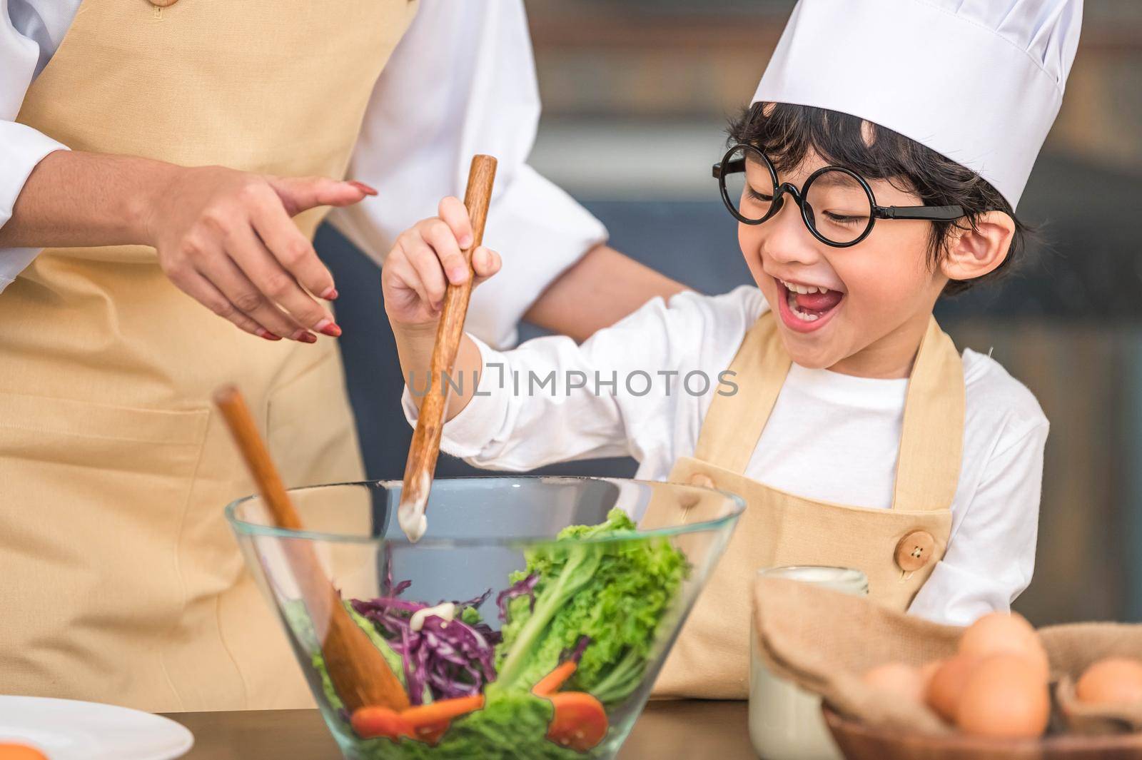 Cute little Asian happy boy interested in cooking with mother funny in home kitchen. People lifestyles and Family. Homemade food and ingredients concept. Two Thai people making ketogenic diet salad