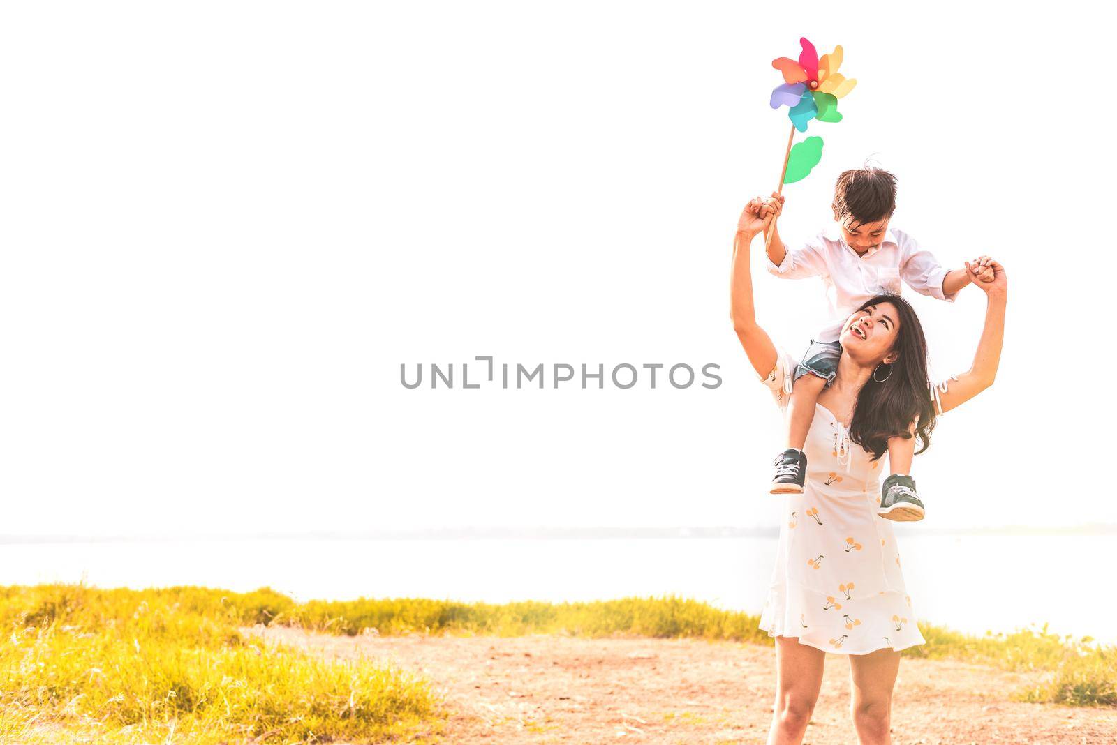 Little Asian boy riding back on his super power mom in meadow near lake. Mother and son playing together. Celebrating in Mother day and appreciating concept. Summer people and lifestyle theme.